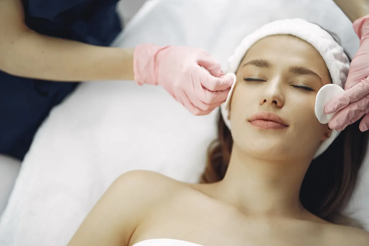 An aesthetician in pink gloves cleansing a relaxed client's face with cotton pads