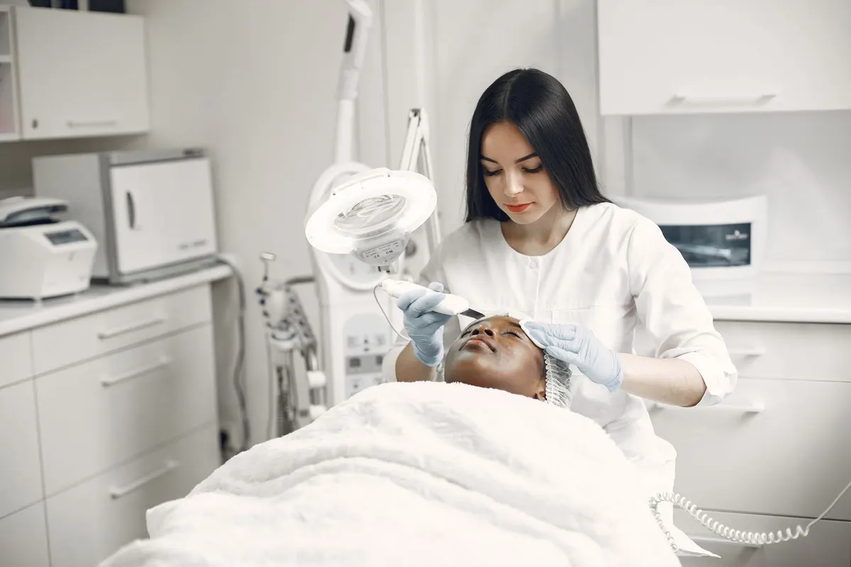 An aesthetician in white giving a client a facial under a magnifier lamp