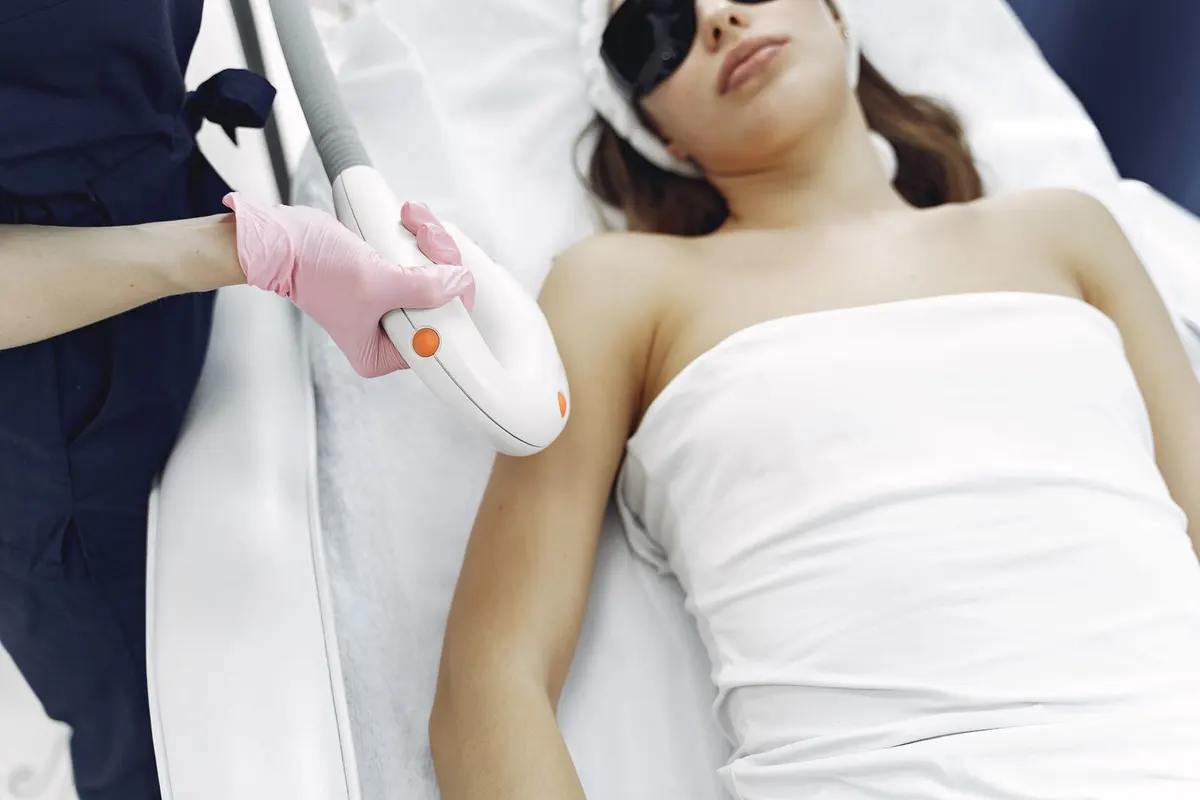A gloved hand holding a laser handpiece over a client's arm while she wears protective glasses
